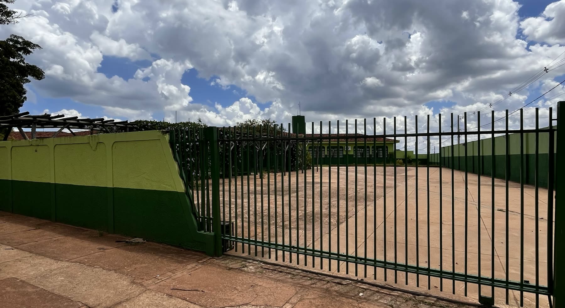 Portón y reja verdes frente a un patio de cemento vacío, con el bloque de la escuela al fondo bajo un cielo nublado