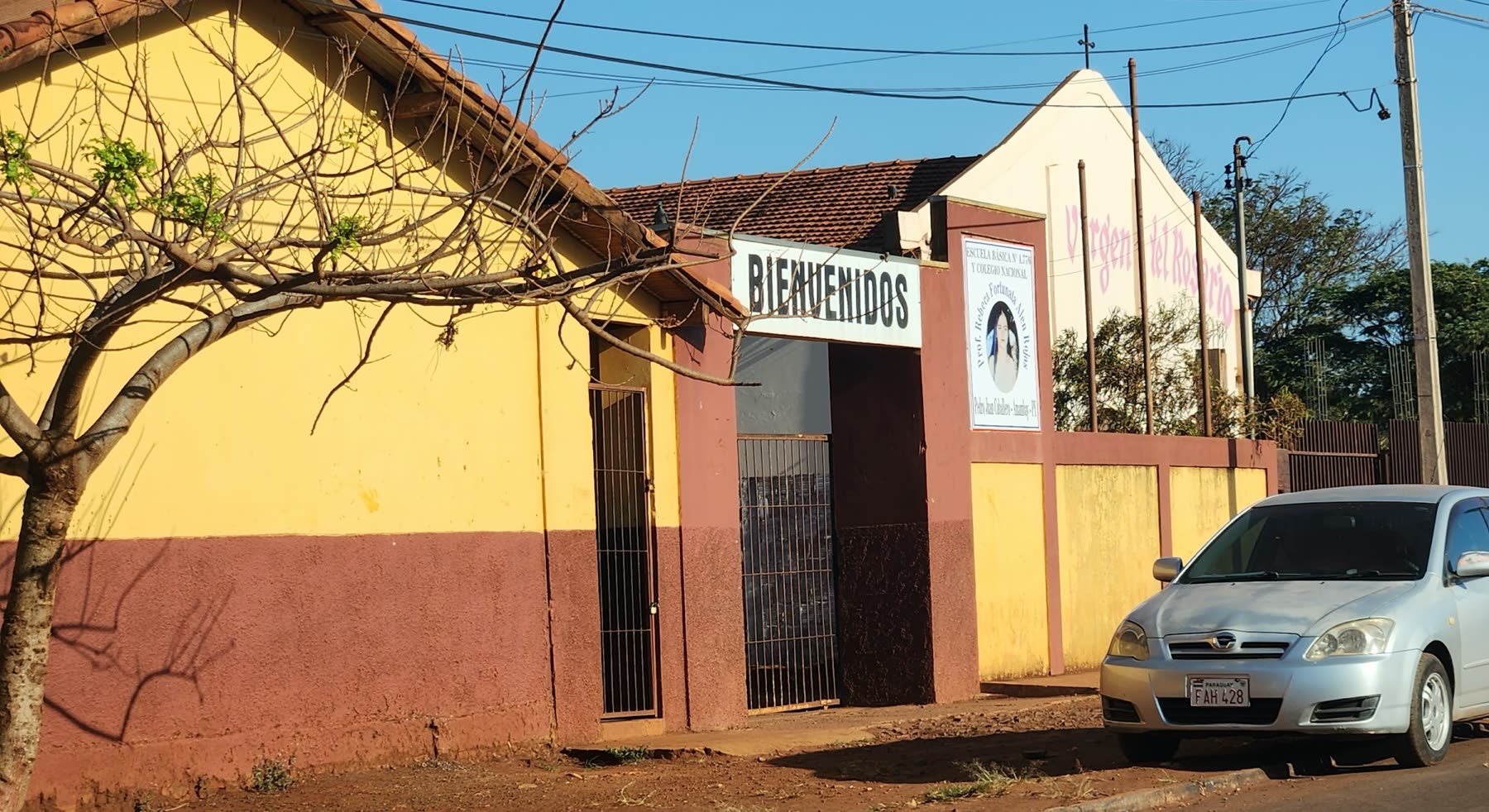 Edificio antiguo de paredes amarillas y terracota, con la palabra Bienvenidos sobre el portón enrejado y un auto detenido en la calle de tierra