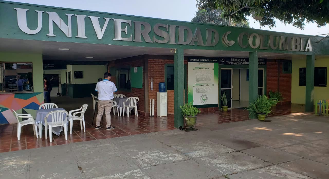 Entrada de edificio verde con las letras Universidad Columbia sobre la marquesina, sillas plásticas blancas y una cartelera de avisos