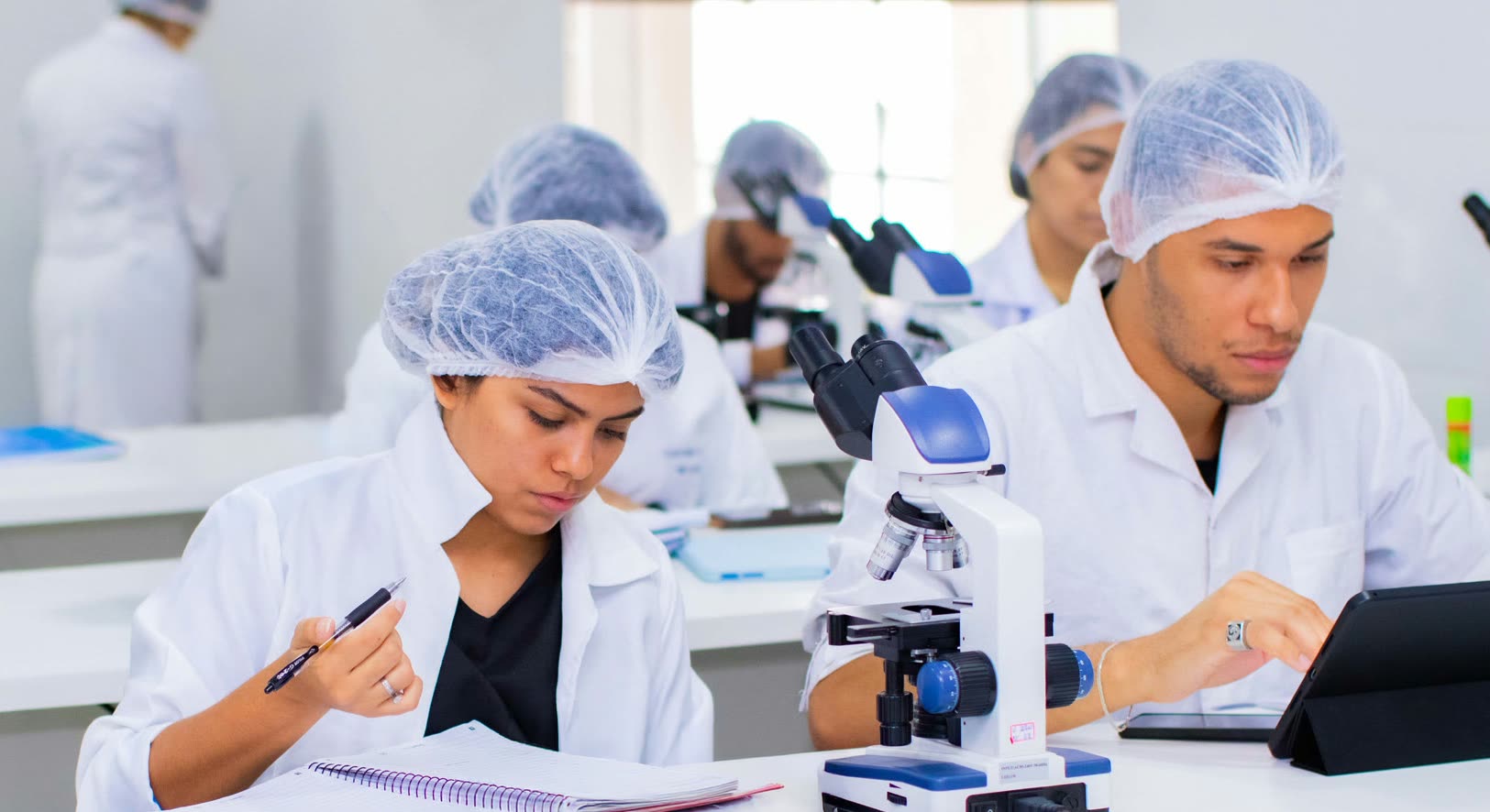 Estudiantes con guardapolvo blanco y cofia descartable en una mesada de laboratorio, con microscopios, un cuaderno abierto y una tableta