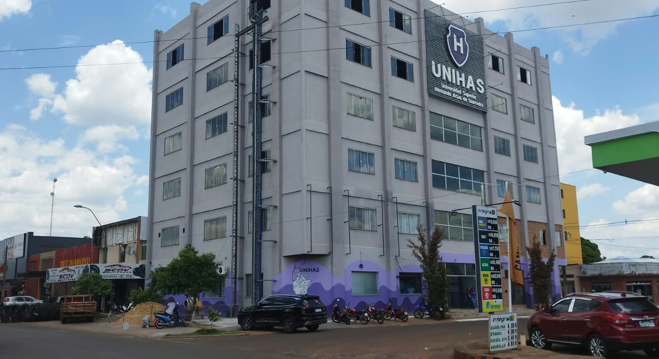 Edificio de cinco pisos en gris y violeta con el escudo de Unihas en lo alto, motos y autos estacionados en la esquina