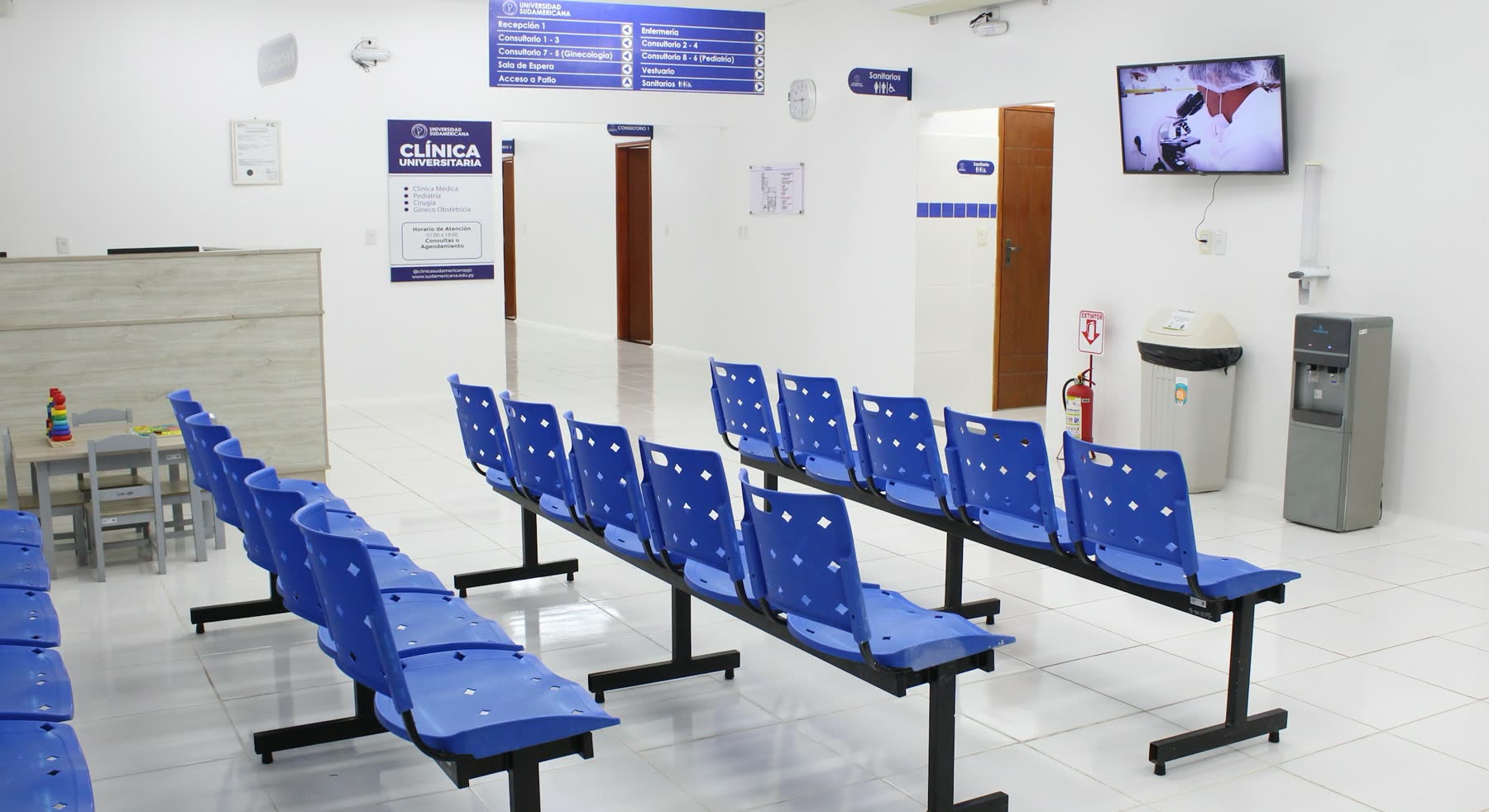 Sala de espera con hileras de sillas plásticas azules, televisor en la pared, dispensador de agua y cartel de señalización de la clínica