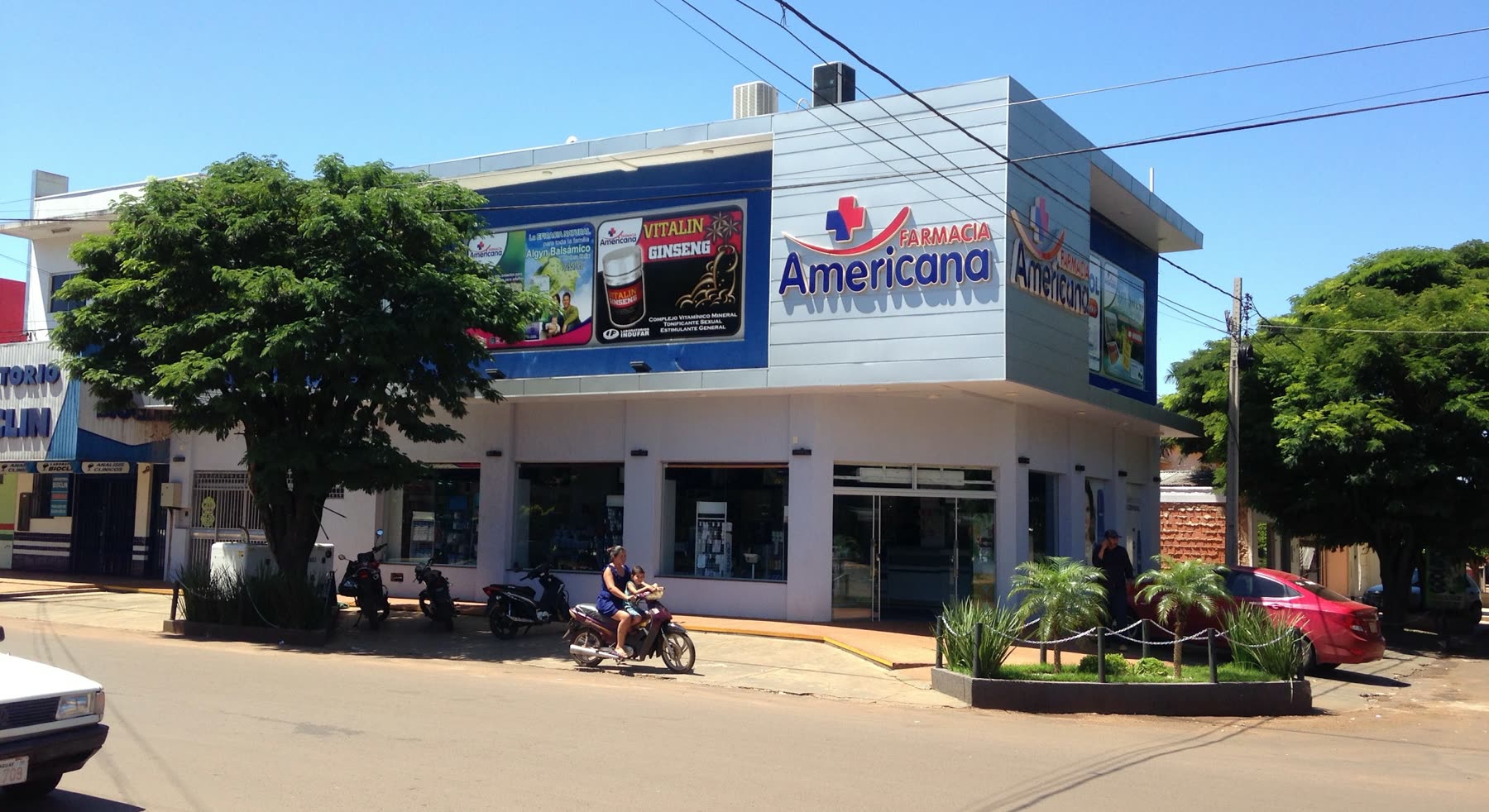 Prédio de esquina azul e branco com o letreiro Farmacia Americana, cartazes de produtos na fachada e uma moto passando na rua