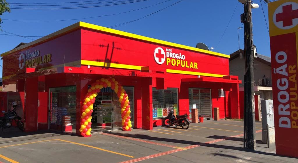 Loja de esquina vermelha e amarela com o letreiro Drogão Popular e um arco de balões na porta de entrada