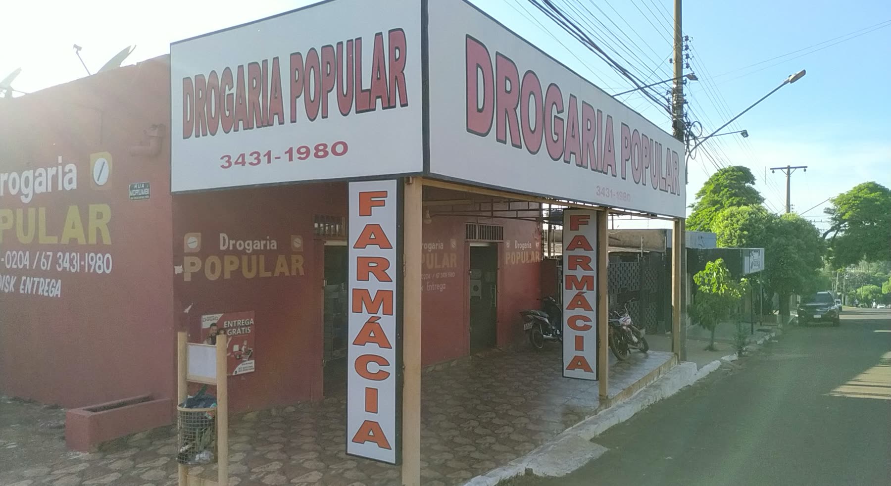 Fachada branca e vermelha com dois letreiros Drogaria Popular e faixas verticais escritas Farmácia