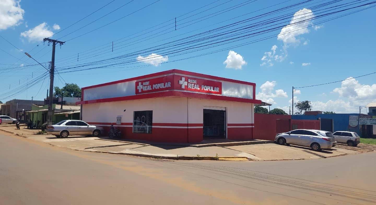 Loja térrea branca e vermelha de esquina com o letreiro Real Popular, carros estacionados na rua de terra e céu de nuvens