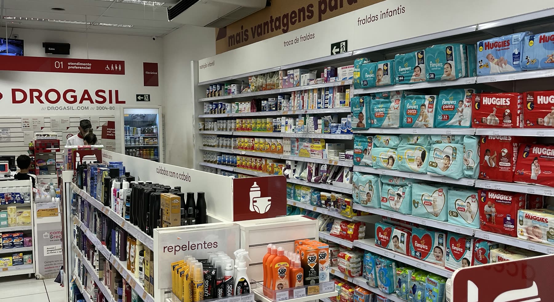 Interior de farmácia com gôndolas de fraldas, repelentes e cosméticos e o letreiro Drogasil acima do caixa