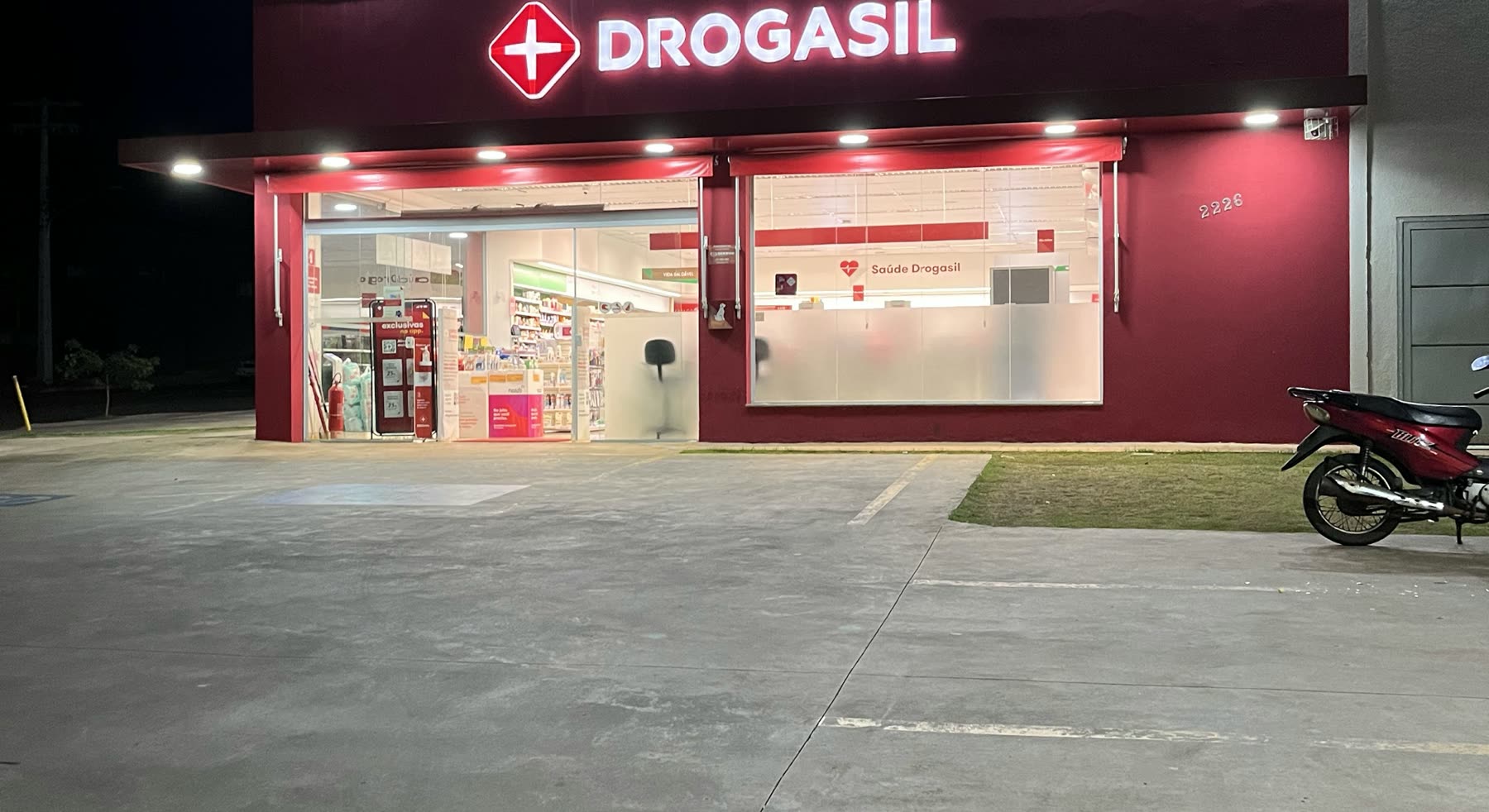 Fachada de vidro iluminada à noite com o letreiro vermelho Drogasil e uma moto estacionada ao lado