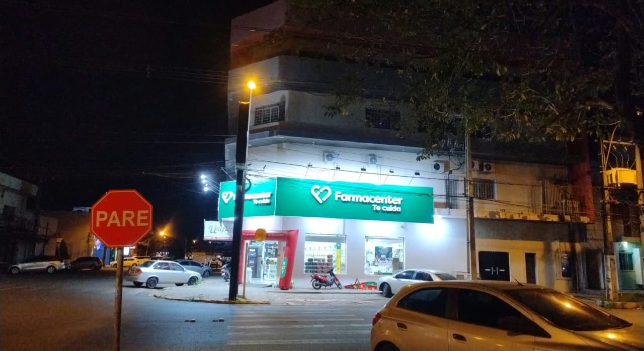 Esquina à noite com o letreiro verde Farmacenter aceso, placa de PARE e carros parados na rua