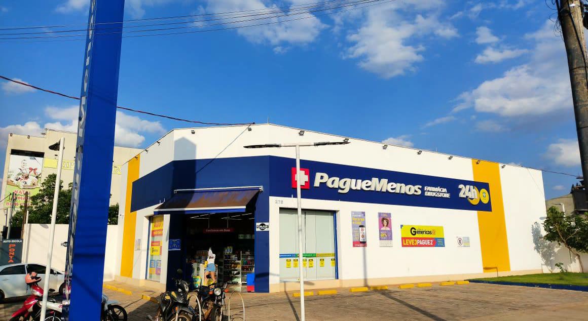 Loja de esquina branca, azul e amarela com o letreiro PagueMenos e o selo de 24 horas, com motos estacionadas na calçada