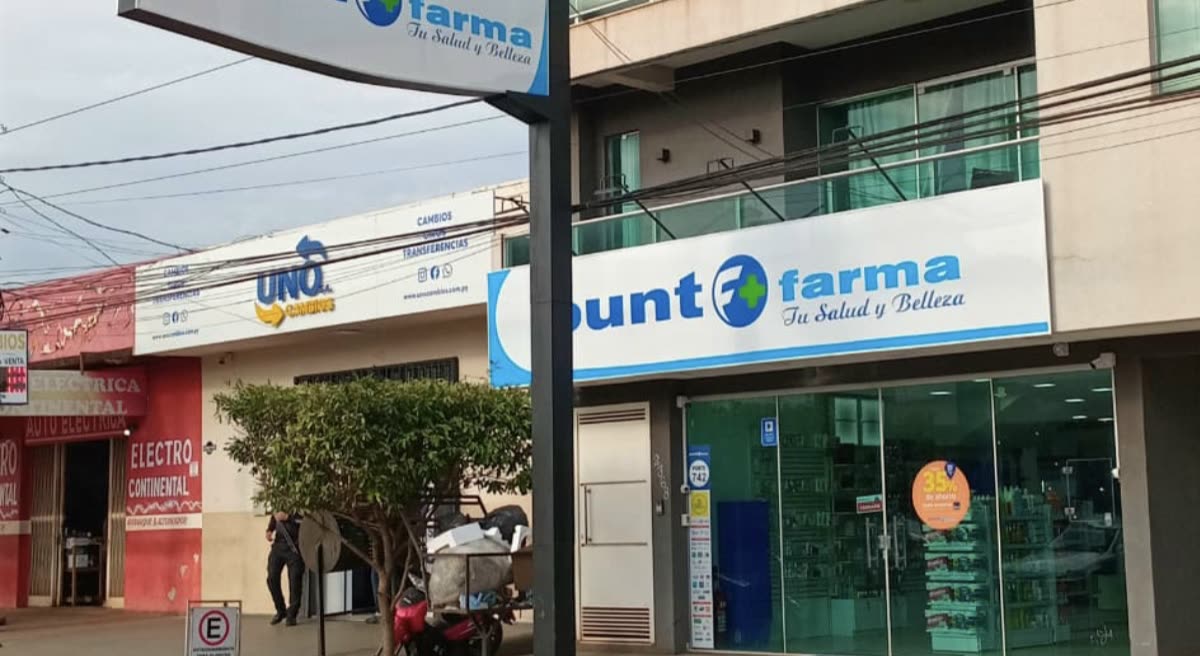 Letreiro azul e branco Punto Farma sobre uma loja envidraçada, com um totem alto na calçada e comércios vizinhos