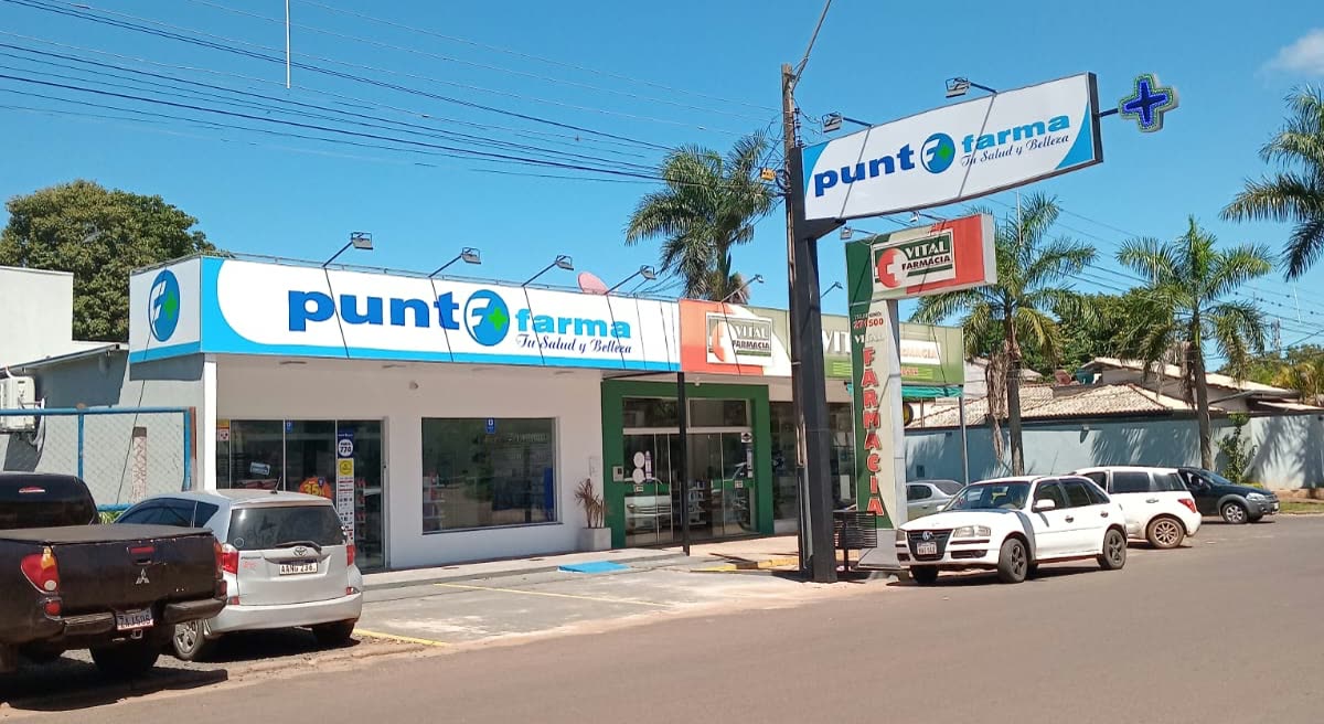 Loja térrea com o letreiro Punto Farma e totem alto na esquina, palmeiras e carros estacionados sob céu azul