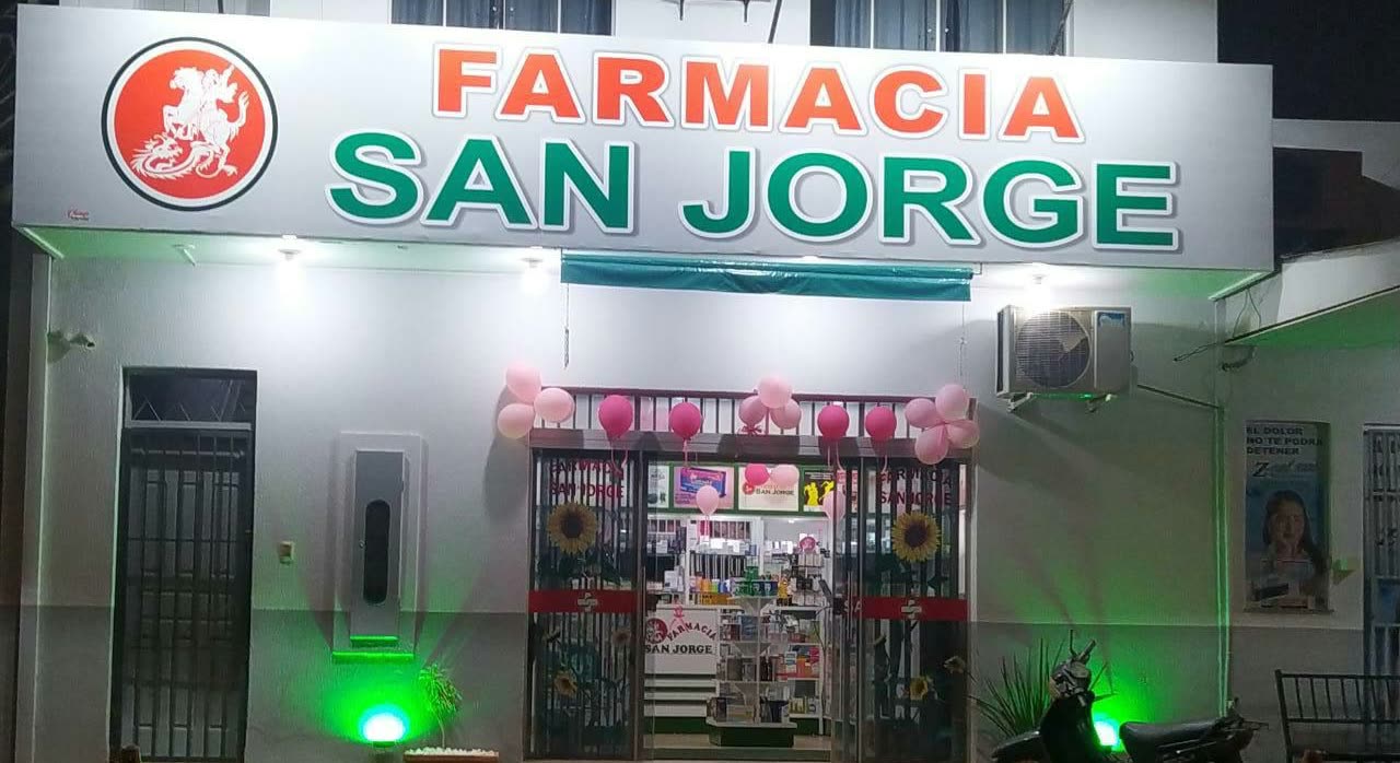 Fachada branca iluminada à noite com o letreiro Farmacia San Jorge e balões cor-de-rosa em volta da porta de vidro