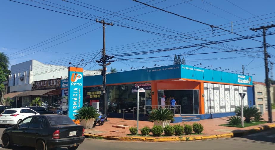 Prédio de esquina em azul e laranja com um totem vertical escrito Farmácia, sob fios de energia e céu limpo