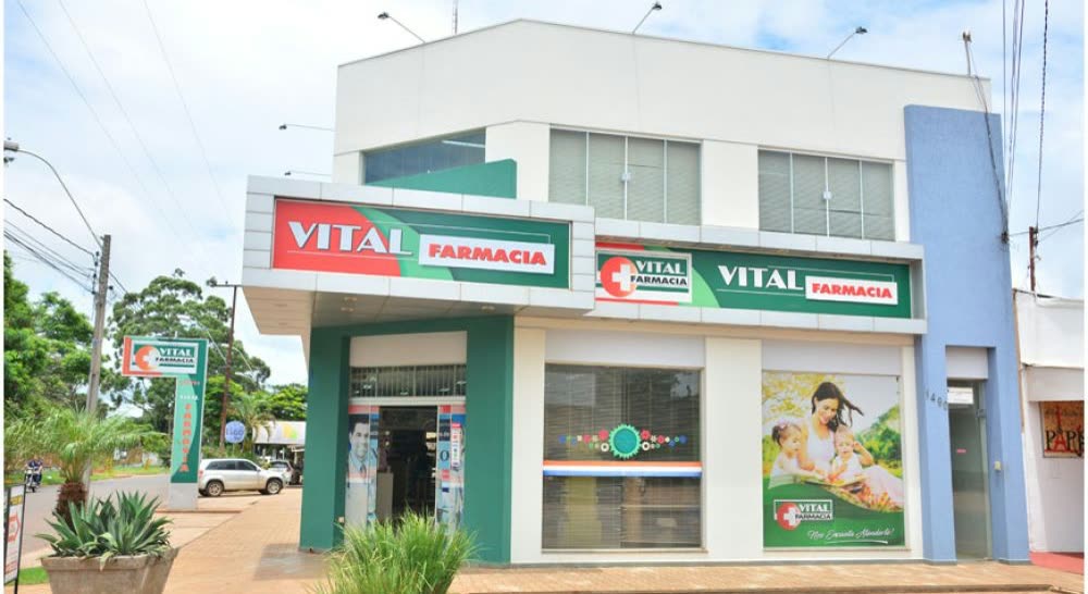 Prédio de dois andares em branco e verde com dois letreiros Vital Farmácia e um painel fotográfico ao lado da porta