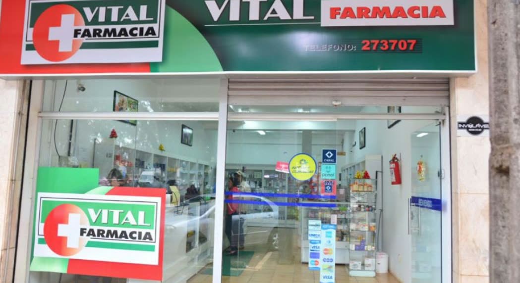 Loja envidraçada sob o letreiro verde Vital Farmacia com o telefone impresso, e as prateleiras visíveis lá dentro