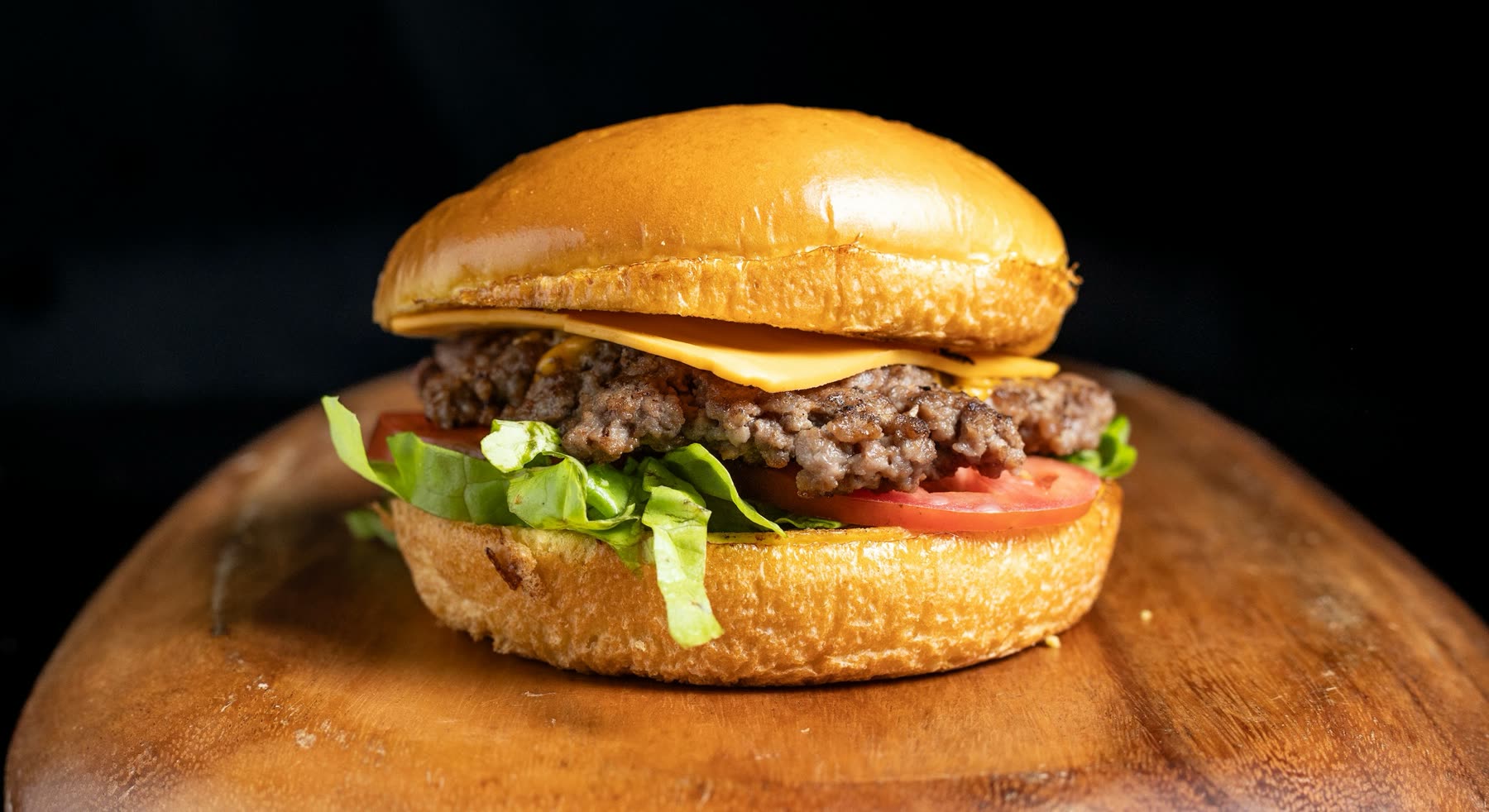 Hamburguesa con queso derretido, lechuga y tomate en pan brioche, sobre tabla redonda de madera y fondo negro