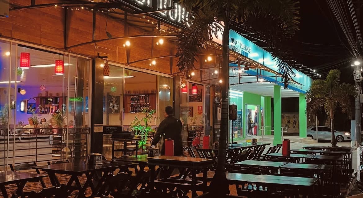 Terraza de noche con mesas y bancos de madera bajo una guirnalda de luces, y una persona de pie junto al mostrador vidriado