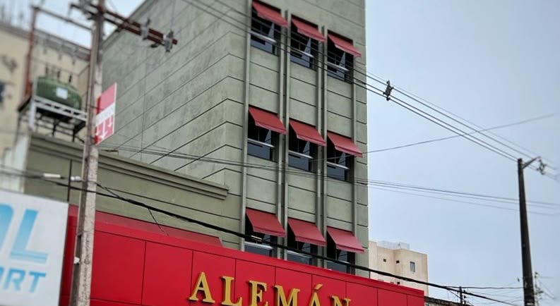 Edificio alto de hormigón con toldos rojos en las ventanas y un panel rojo con las letras doradas Alemán a nivel de la calle