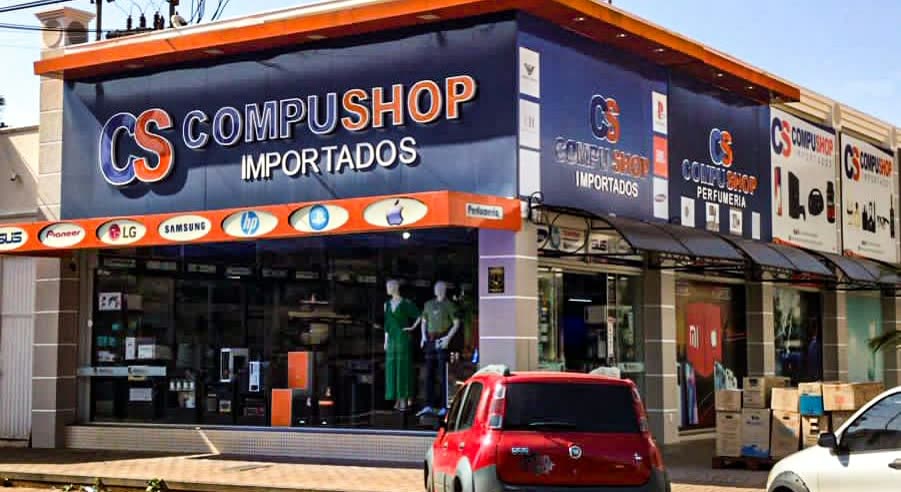 Local de esquina en naranja y azul con el letrero CompuShop Importados, logotipos de marcas de electrónica en la vidriera y un auto rojo estacionado enfrente
