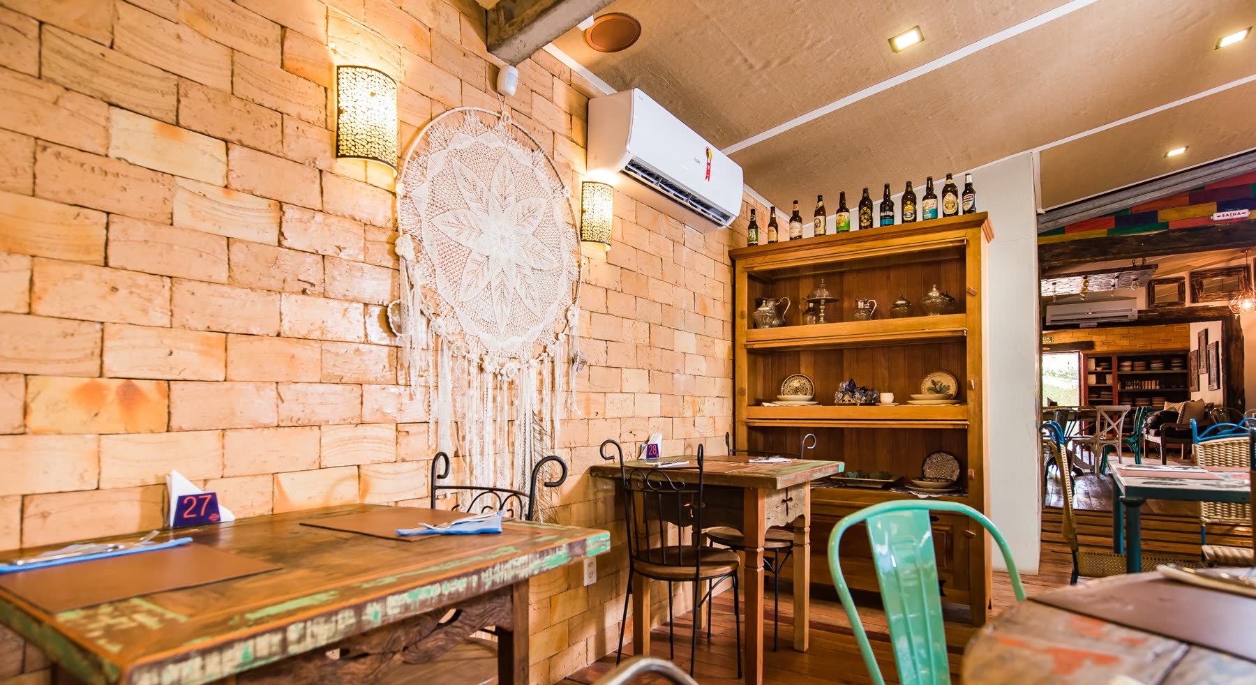 Salón de pared de ladrillo a la vista con un atrapasueños grande, estantería de botellas, mesas de madera y sillas de metal