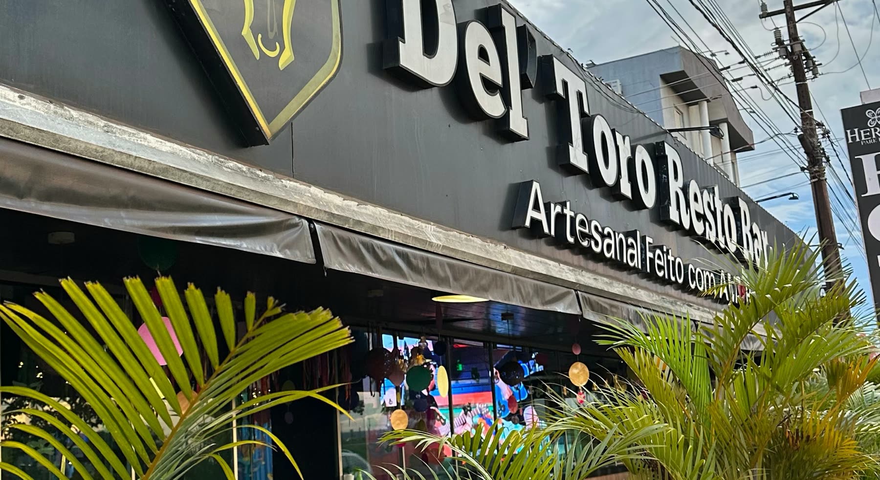 Letrero negro con las letras Del Toro Resto Bar y la frase Artesanal Feito com Amor, visto detrás de hojas de palmera