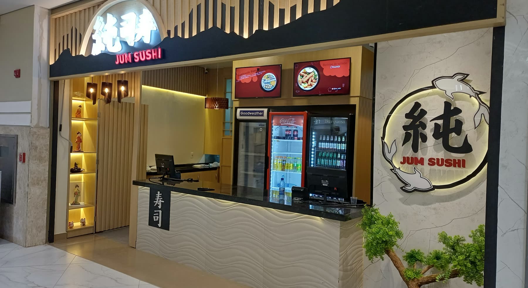 Puesto de shopping con el letrero Jum Sushi en ideograma, mostrador de atención, heladeras de bebidas y un bonsái al costado