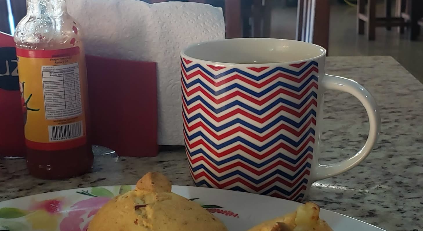 Taza rayada en rojo, azul y blanco junto a una botella y un plato con chipas, sobre una mesa de granito