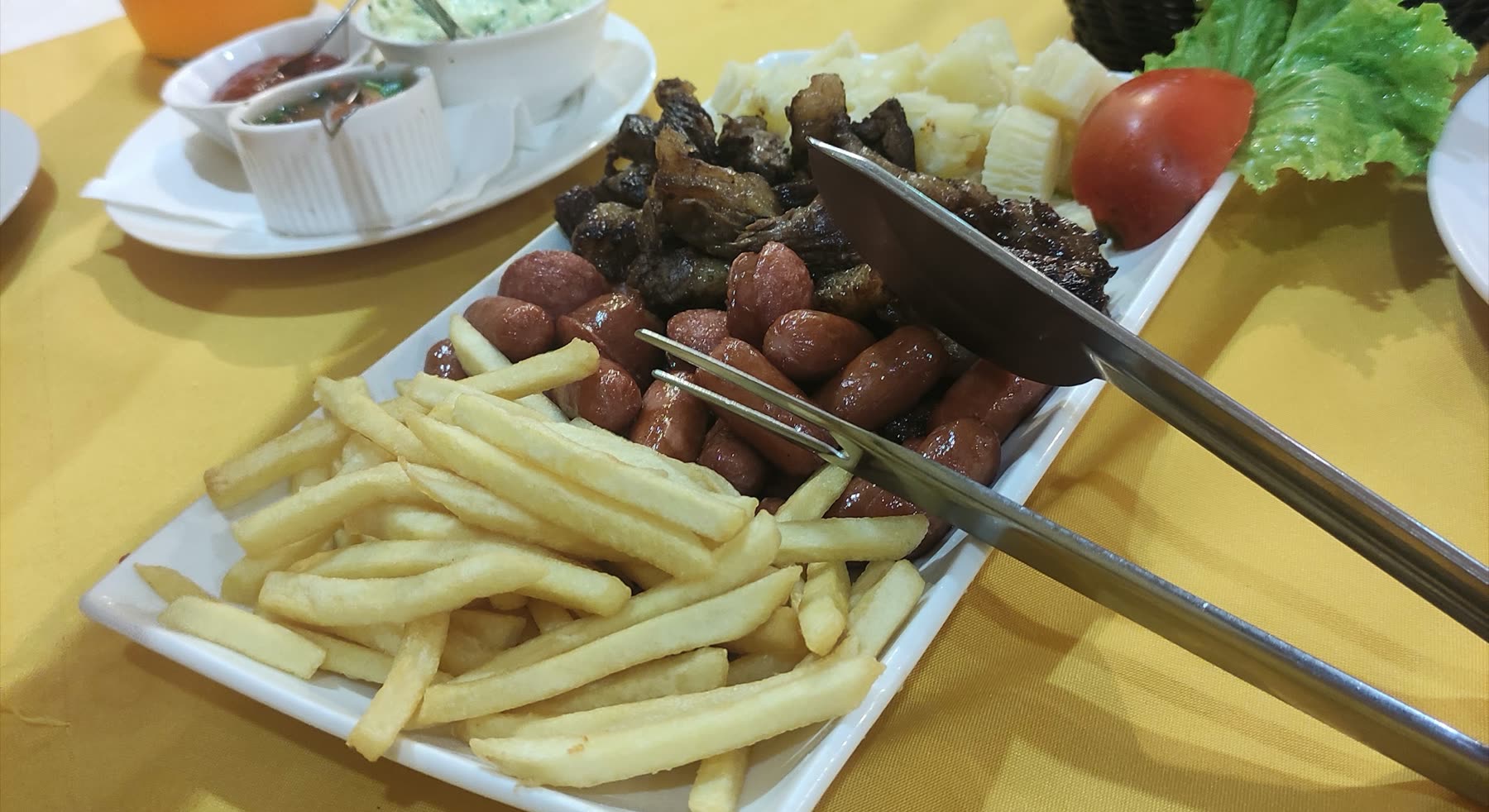 Fuente con papas fritas, chorizo, trozos de carne asada y mandioca, con pinza, sobre mantel amarillo