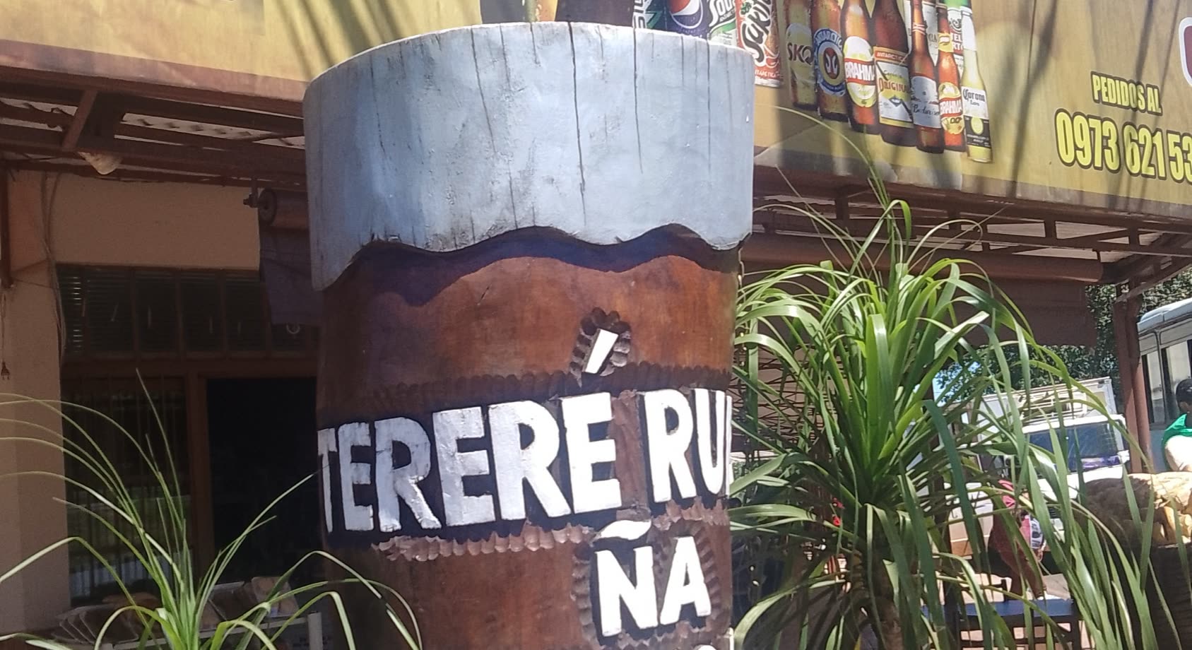 Tótem de madera con forma de guampa gigante con las palabras Tereré Ru y Ña pintadas, rodeado de plantas