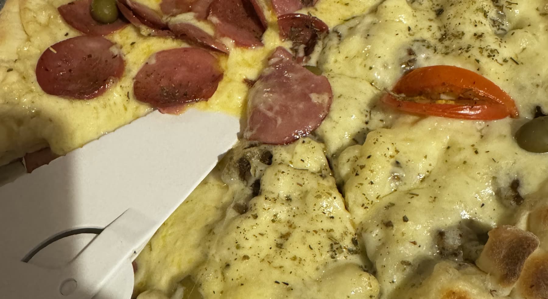 Pizza en primer plano con fetas de salame, aceitunas, tomate, queso derretido y orégano, con una espátula de plástico apoyada