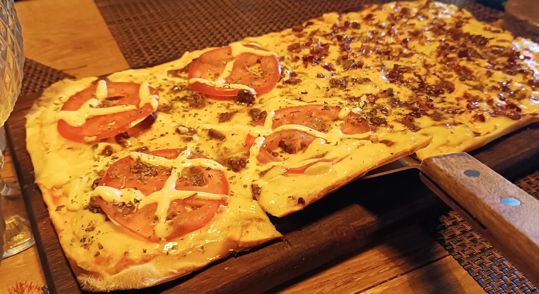 Pizza rectangular con rodajas de tomate, hilos de mayonesa y cobertura granulada, servida sobre tabla de madera