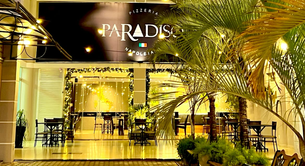 Fachada negra iluminada con el letrero Pizzeria Paradiso, mesas altas al frente y palmeras en macetas