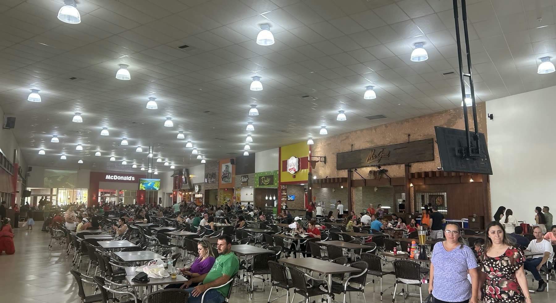 Patio de comidas repleto, con hileras de mesas y sillas bajo el techo de luminarias y locales al fondo